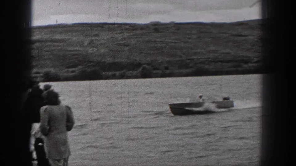 Home-movie frame from 1938 Washington showing distant shore viewers beside the water while a small open boat passes in the background.