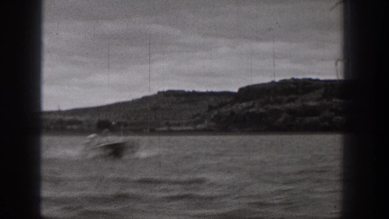 Black-and-white Washington home-movie frame of a small blurred boat crossing choppy water below a low hillside shoreline.