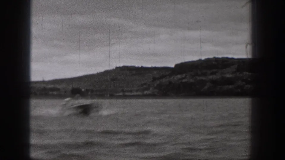 Black-and-white Washington home-movie frame of a small blurred boat crossing choppy water below a low hillside shoreline.