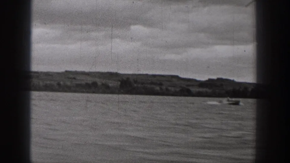 1938 Washington 8mm frame showing a small boat far out on calm water, with a low shoreline and overcast sky beyond dark film borders.