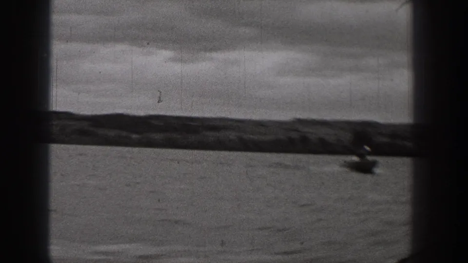 1938 Washington home-movie frame with a small distant boat near the right edge, calm water, low shoreline, and heavy clouds.