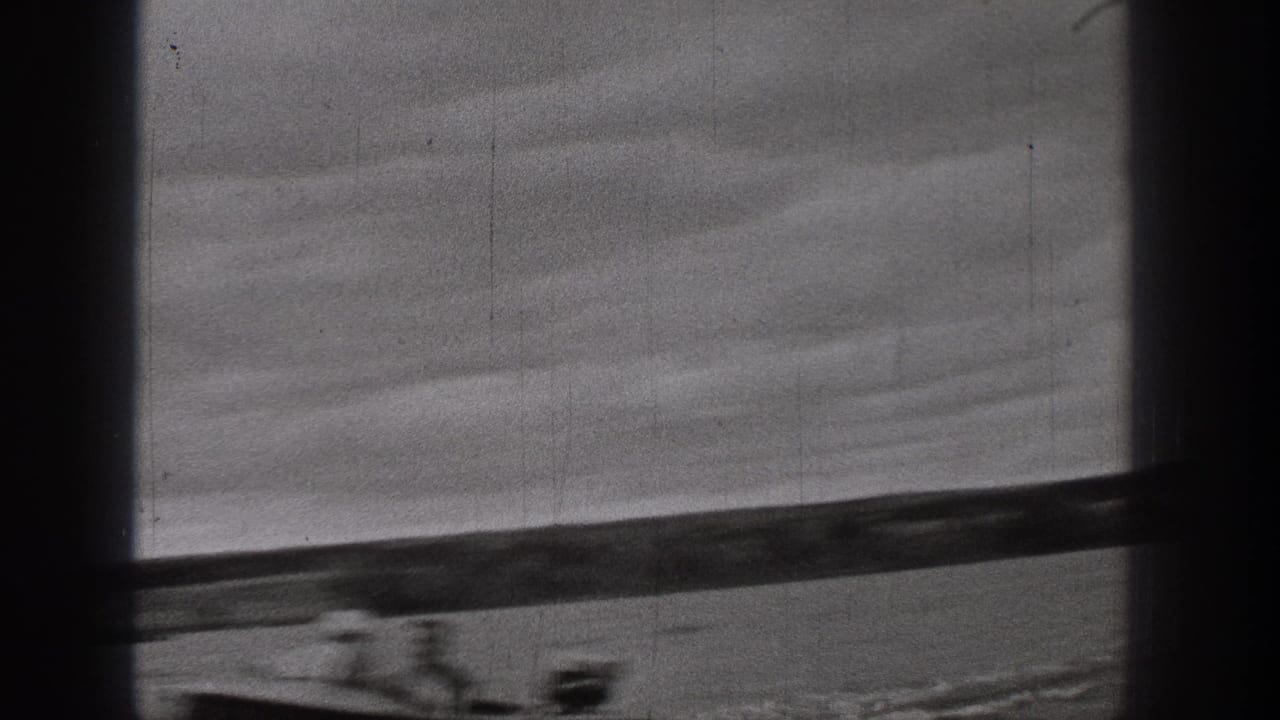 1938 Washington home-movie frame with calm water, low shoreline, heavy cloud cover, dark borders, and a faint boat wake near the bottom edge.