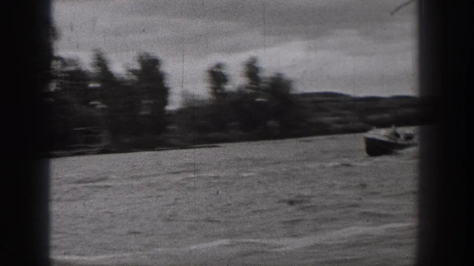 Small motorboat crosses a Washington lake in soft black-and-white 8mm footage, with trees and low shoreline in the background.