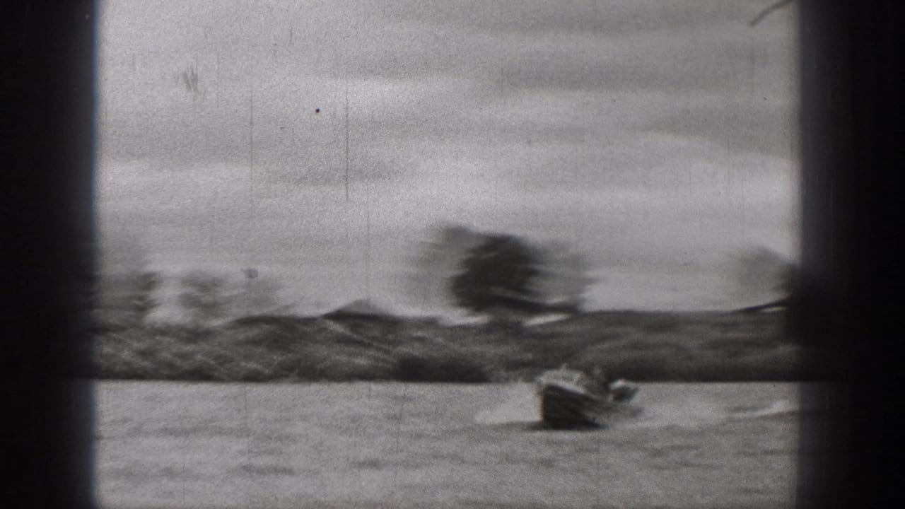Black-and-white 8mm frame of a small motorboat on a Washington lake, with low hills, scattered trees, and cloudy sky beyond.