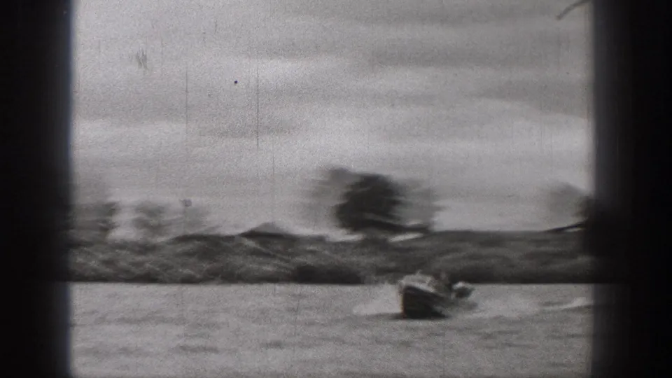 Black-and-white 8mm frame of a small motorboat on a Washington lake, with low hills, scattered trees, and cloudy sky beyond.
