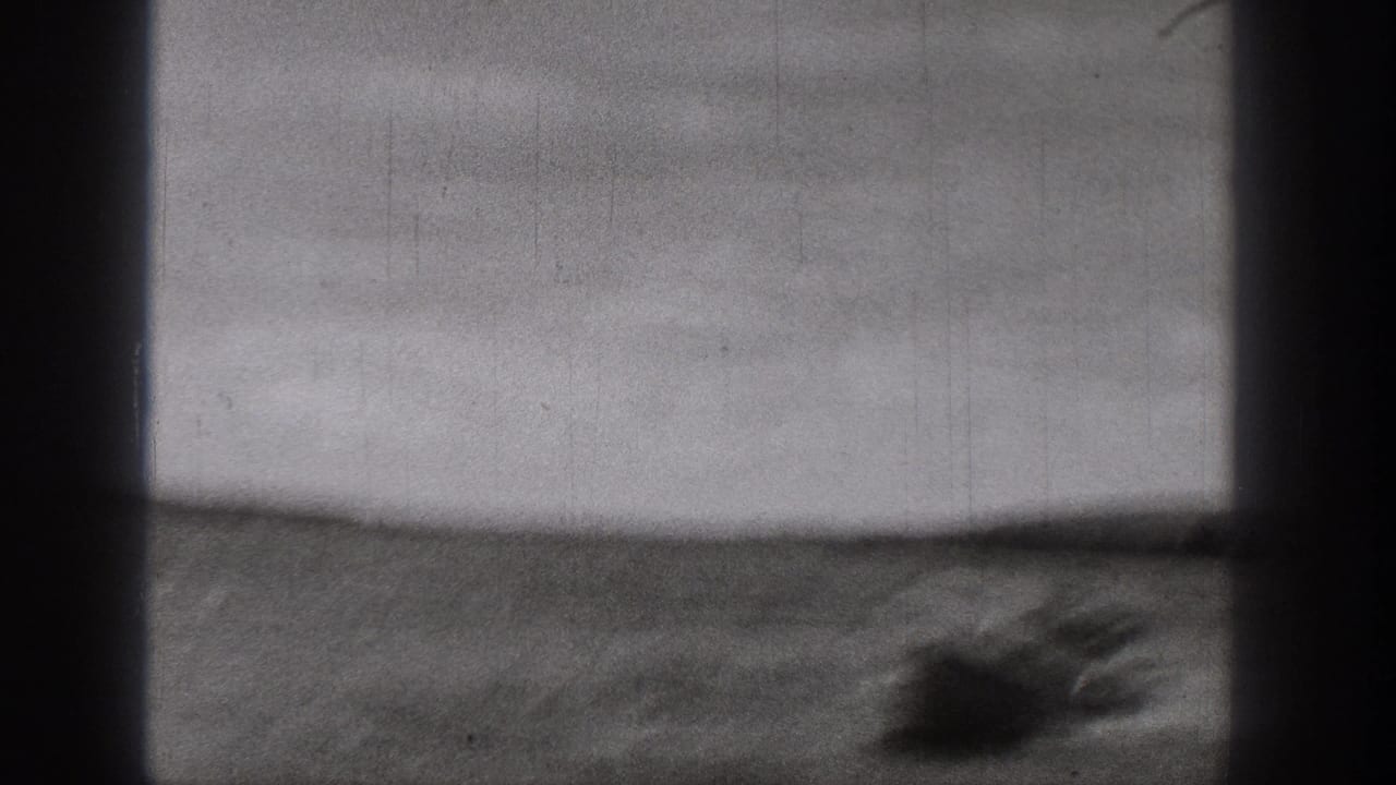 Black-and-white 8mm frame showing a small boat near the lower right, open water, low shoreline, and soft film grain.