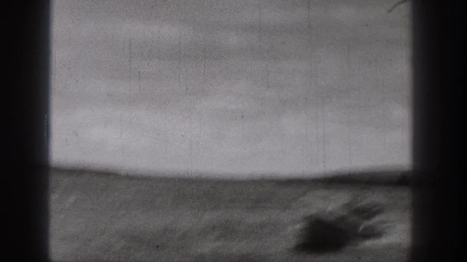 Black-and-white 8mm frame showing a small boat near the lower right, open water, low shoreline, and soft film grain.