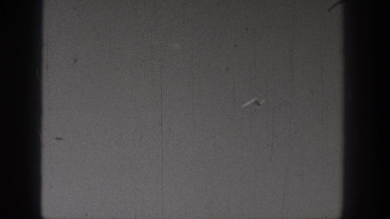 Soft black-and-white 8mm frame with a tiny pale model-plane shape against a mostly empty outdoor background.
