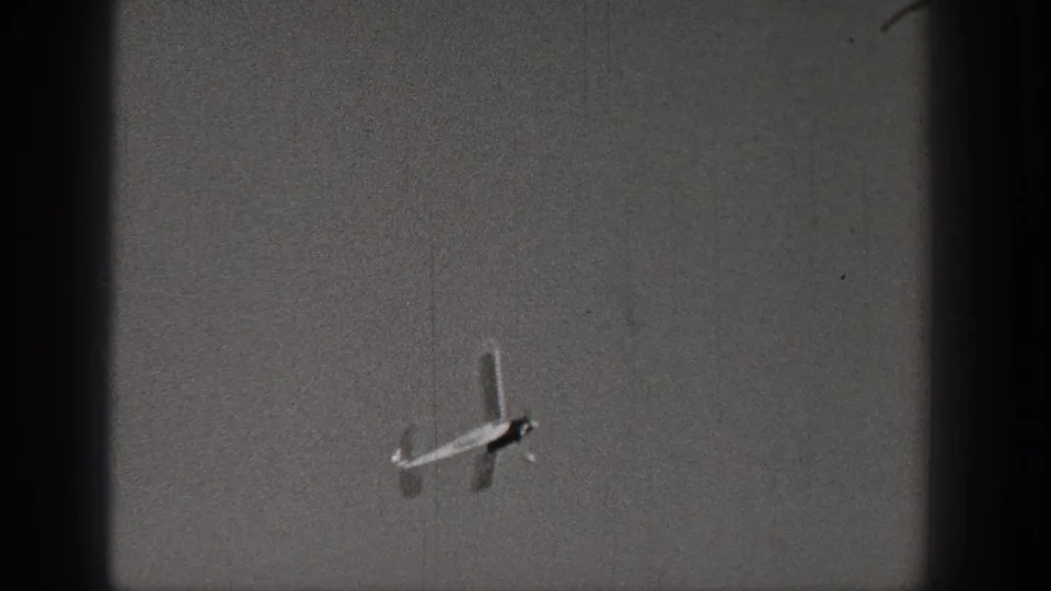 A small model airplane is seen at a distance in the upper sky, framed by 8mm film grain, scratches and dark vertical gate edges.