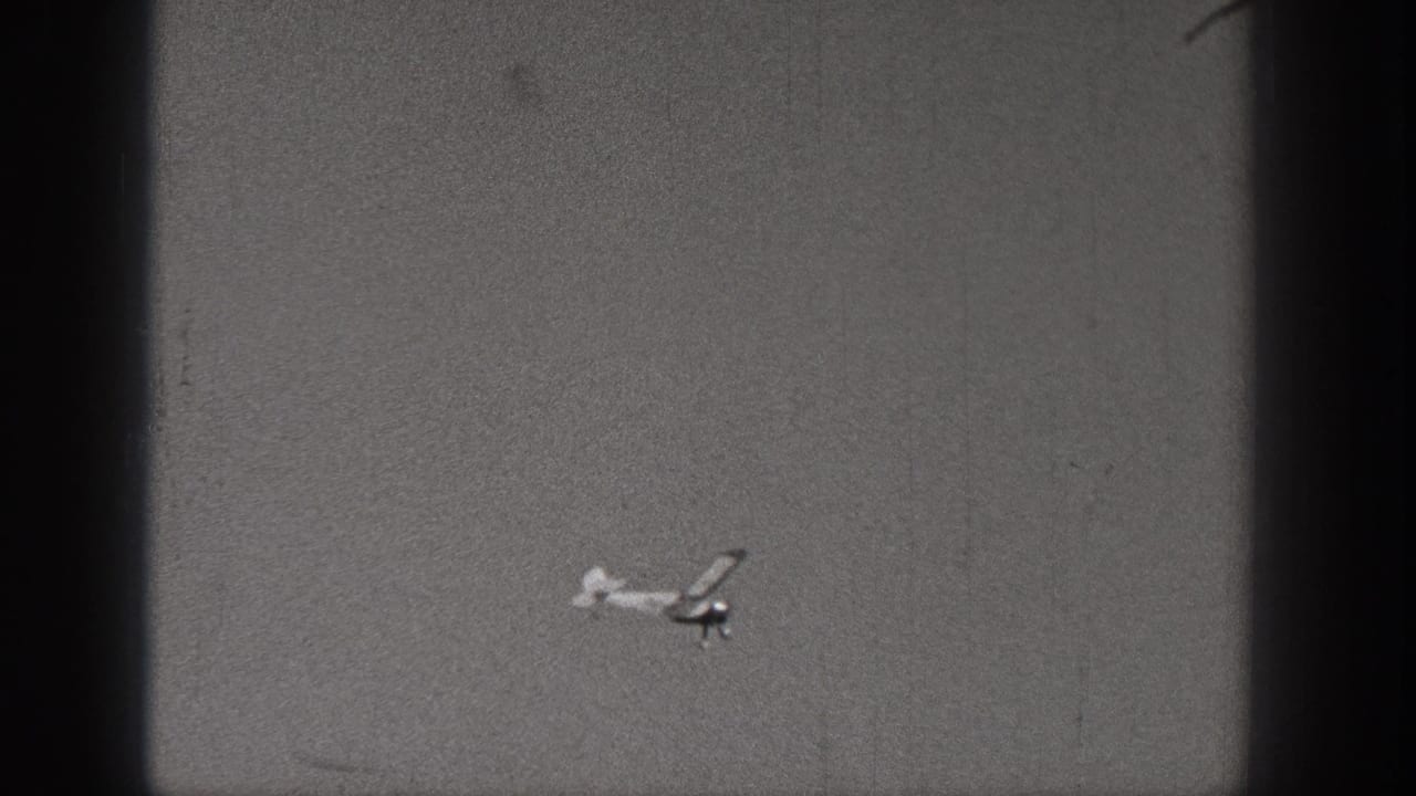 A small model airplane passes low in the frame, its light body and dark shadowed parts visible through grainy 1938 Washington 8mm footage.