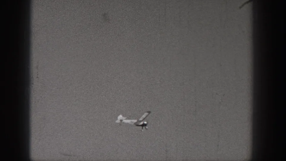 A small model airplane passes low in the frame, its light body and dark shadowed parts visible through grainy 1938 Washington 8mm footage.