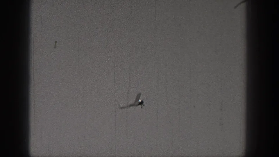 A distant model airplane appears as a darker silhouette against the sky, with visible grain, scratches and the cropped edges of the 8mm frame.