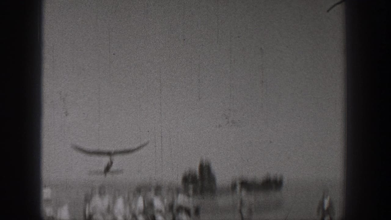 Small glider above a distant group of people in an open field.