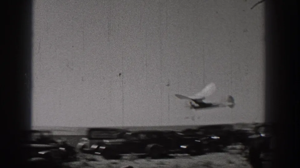 1938 Washington home-movie frame showing the small glider flying low above parked cars and an open field.