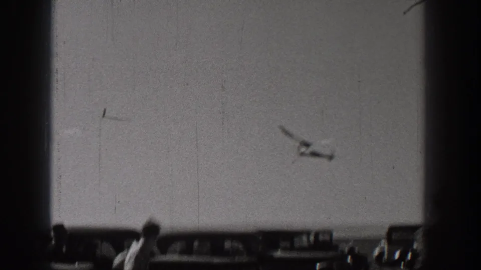 In Washington, United States, 1938, a radio-controlled model airplane appears in flight above parked cars and a few spectators.
