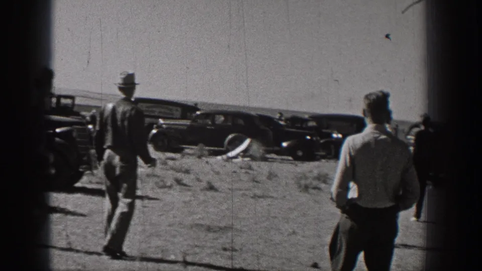 Men in period clothing move across an open field near vintage automobiles during the Washington radio-control plane test.