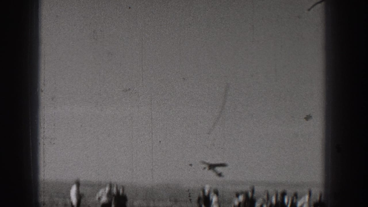A model airplane appears low in the frame over a distant group of spectators, with heavy 8mm grain and scratches visible.