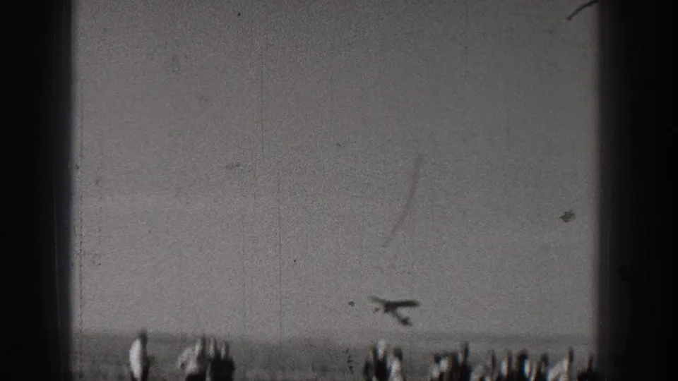 A model airplane appears low in the frame over a distant group of spectators, with heavy 8mm grain and scratches visible.