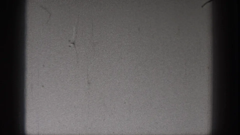 Black-and-white 8mm frame showing open sky within dark gate borders, with scratches and dust but no distinct people or aircraft visible.