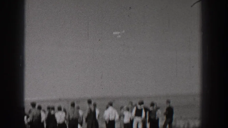 In the supplied 1938 Washington context, spectators in period clothing stand near a shoreline while looking toward activity off frame.