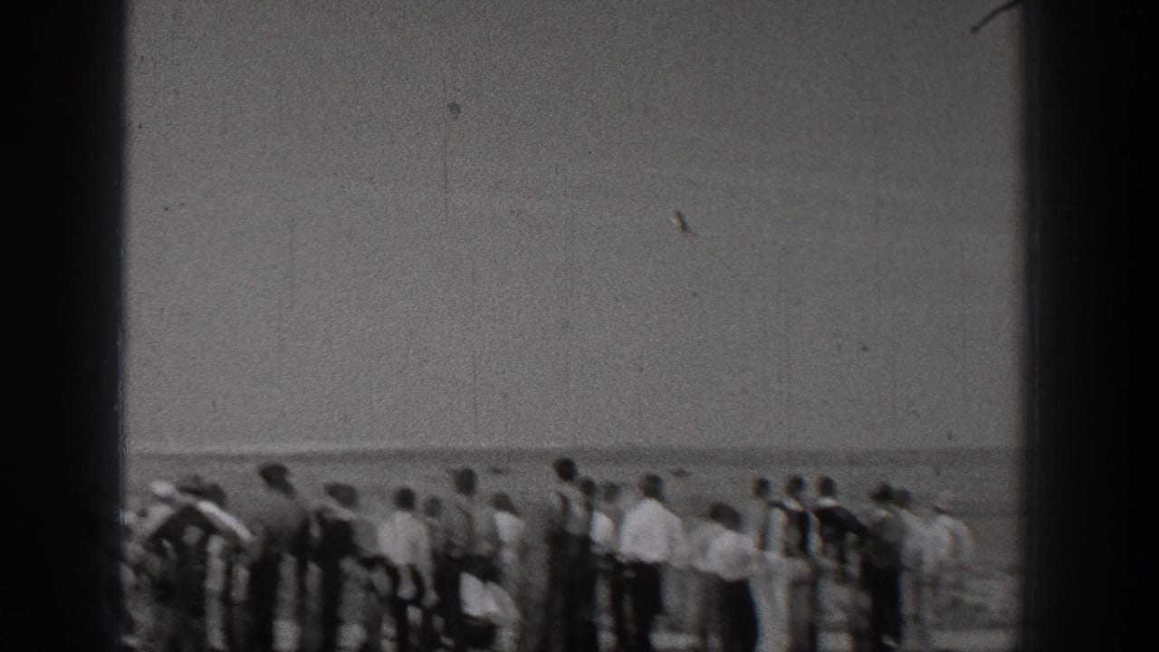 A 1938 Washington home-movie frame shows adults gathered beside the water, watching something beyond the camera's view.