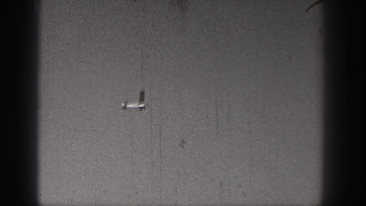 Small light aircraft viewed against a gray sky in a scratched, grainy 1938 Washington 8mm frame.