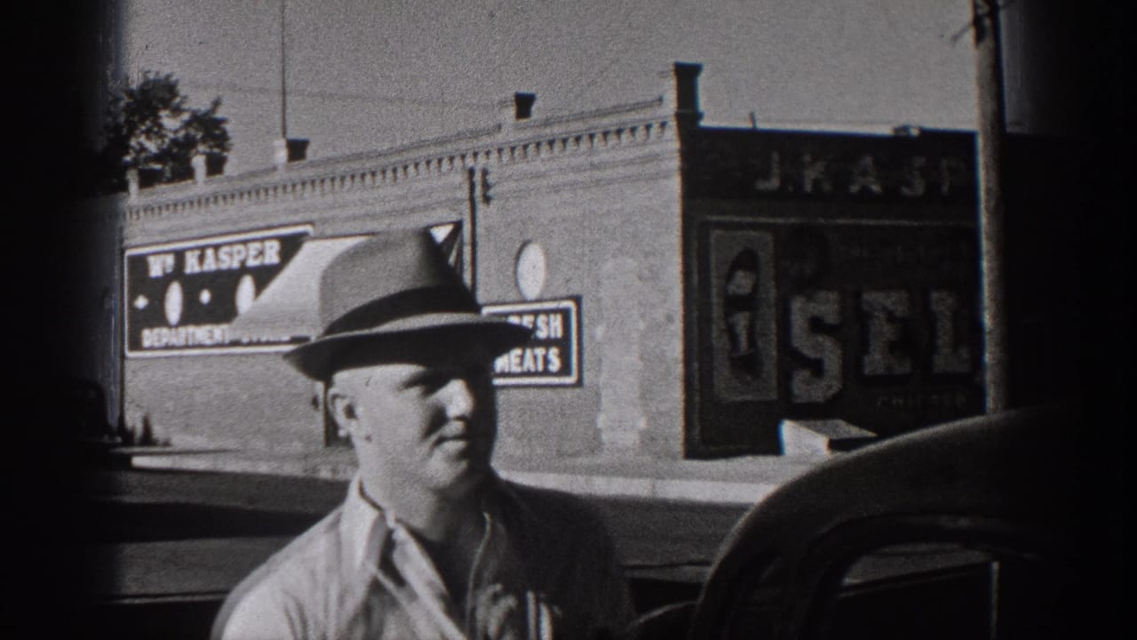 Closer 1938 Washington home-movie frame of a man in a hat before brick storefronts with visible signage and parked cars.