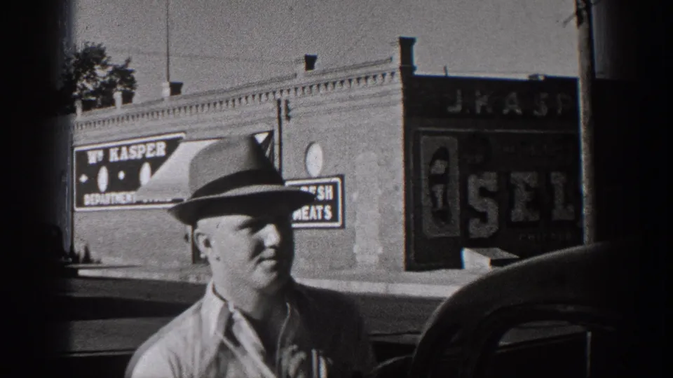 Closer 1938 Washington home-movie frame of a man in a hat before brick storefronts with visible signage and parked cars.