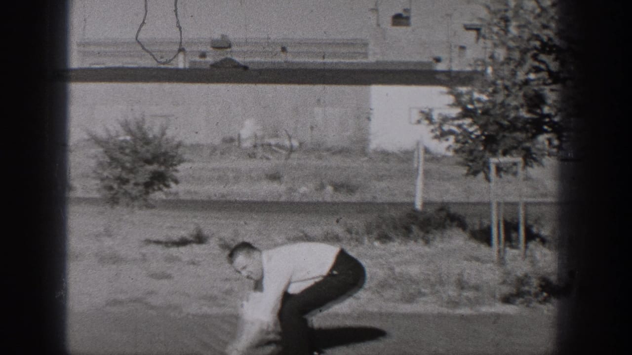 Man in shirt and tie bends down in a grassy roadside area beside a tree and building wall.