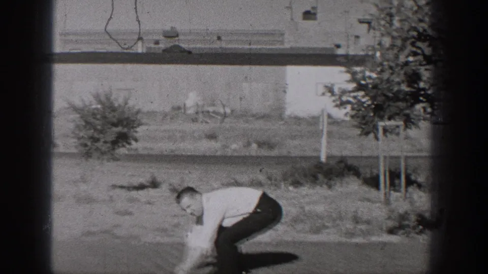 In 1938 Washington footage, a man in a white shirt and tie bends in a grassy lot near a tree and plain building wall.