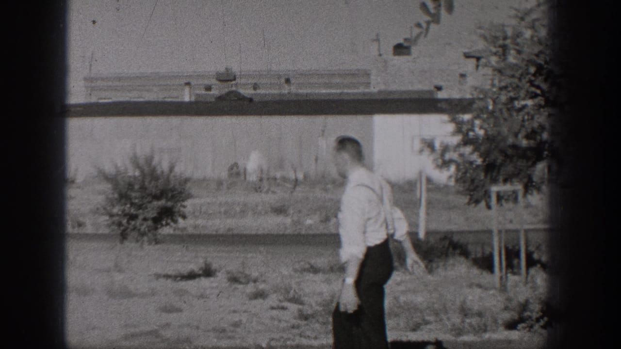 A man in dress clothes walks away from the camera across a grassy Washington roadside area in black-and-white 8mm footage.
