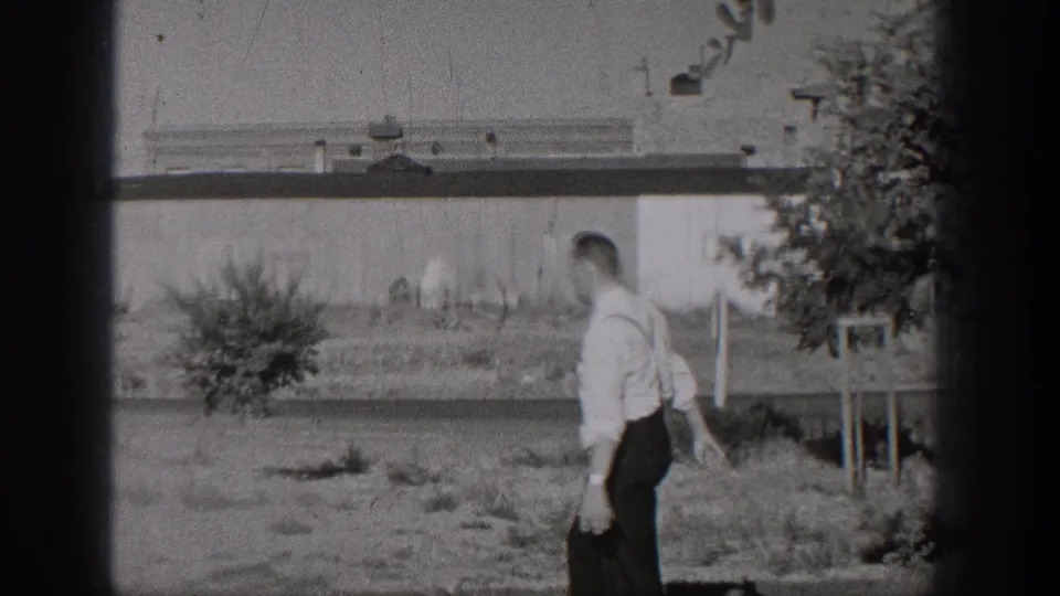 A man in dress clothes walks away from the camera across a grassy Washington roadside area in black-and-white 8mm footage.