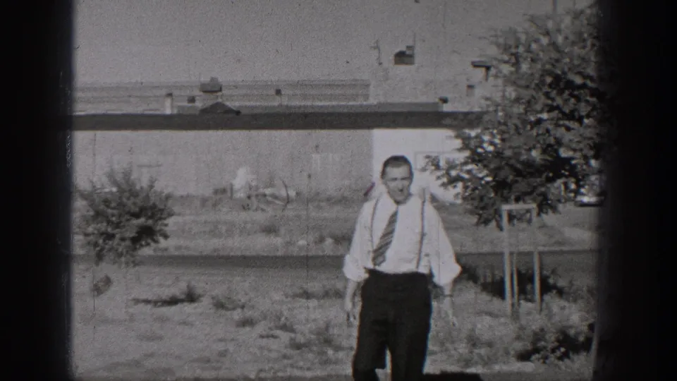 A man in shirt, tie, and suspenders walks toward the camera in a 1938 Washington home-movie street-side scene.