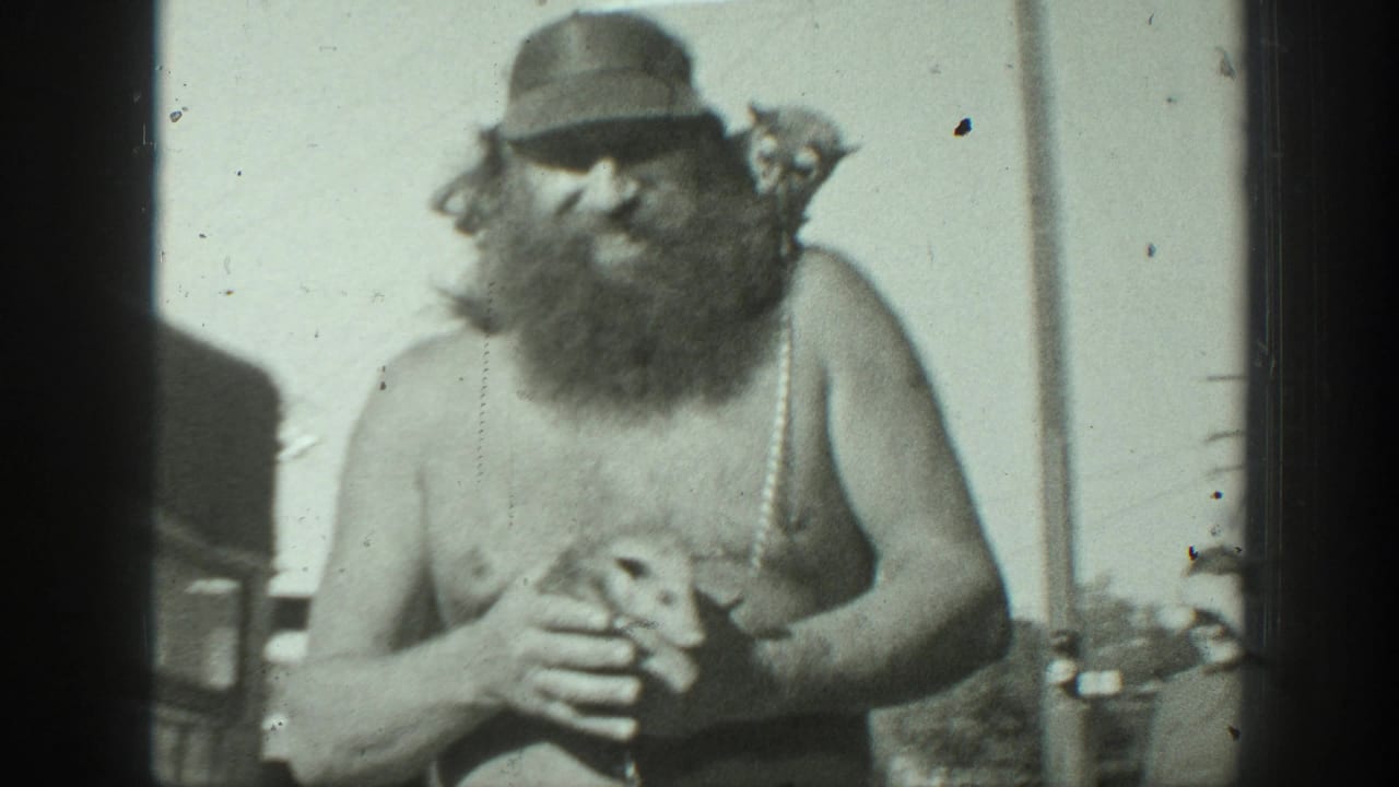 Bearded shirtless man in a cap holding a small animal outdoors.