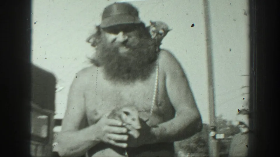 In metadata-supported 1939 California footage, a bearded shirtless man in a cap cradles a small animal outdoors.