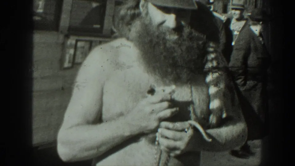 The shirtless bearded man holds the small animal as onlookers pass behind him in the California outdoor setting.
