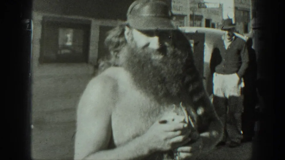 The man turns slightly with the animal in his arms, with pedestrians and a vehicle-like shape in the background.