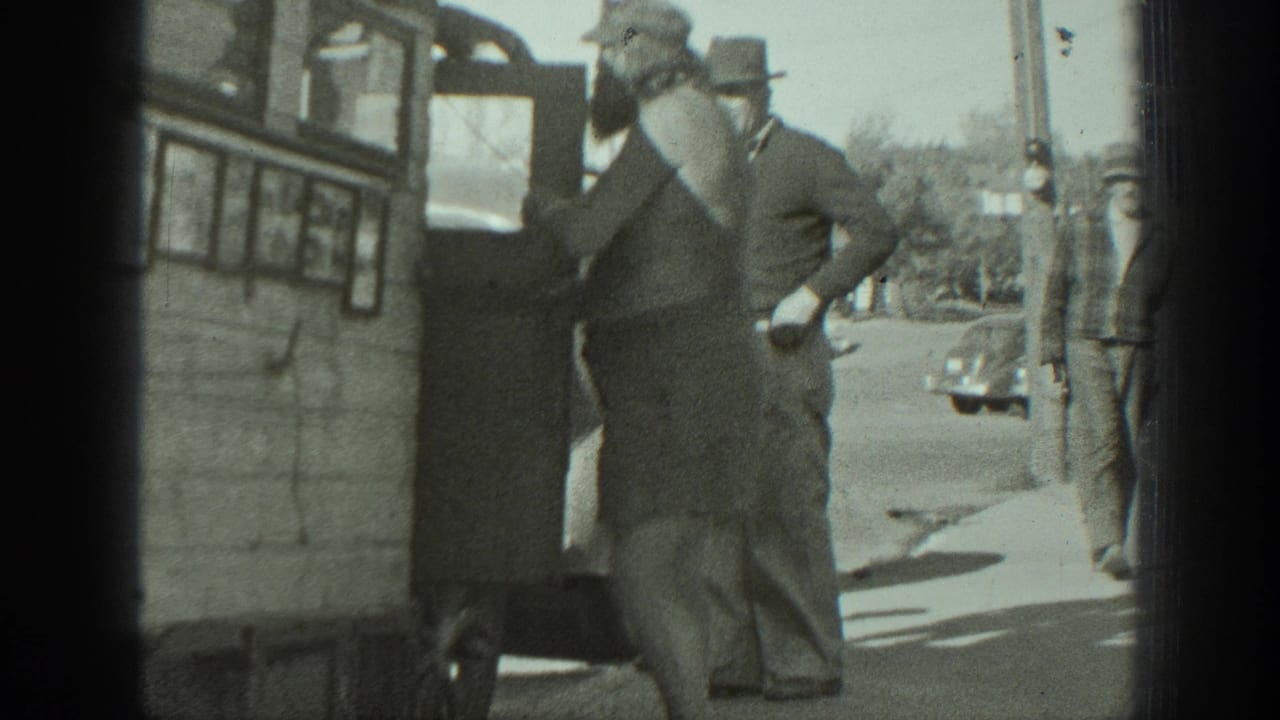 Bearded man holding a small dog beside a bus or camper as men stand on a sidewalk.