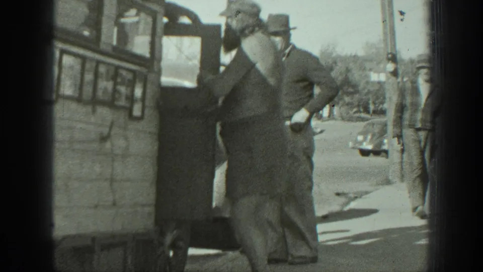 1939 California home-movie frame showing a bearded man carrying a small dog near a bus or camper doorway while men watch from the sidewalk.