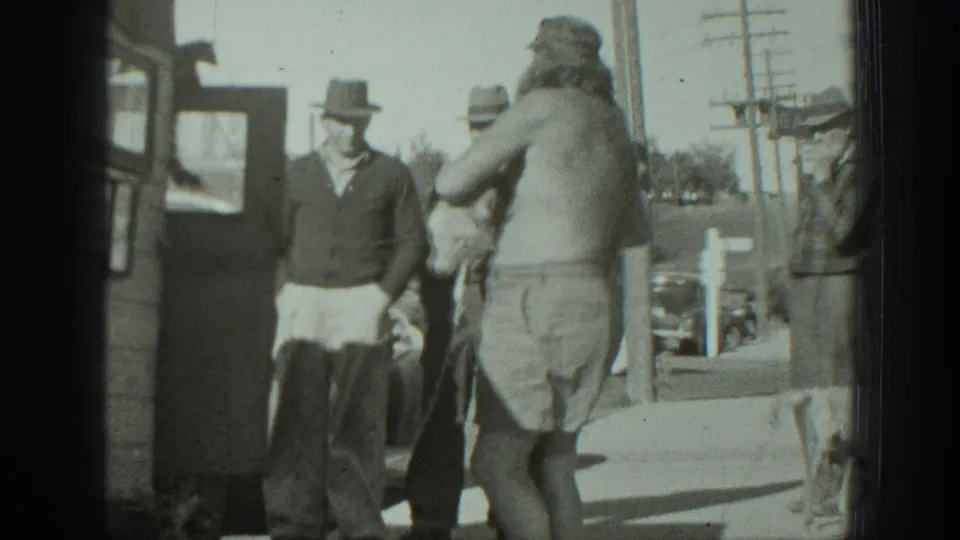 1939 California frame with a bearded man holding a dog near a vehicle, facing men in hats on a sunlit sidewalk.