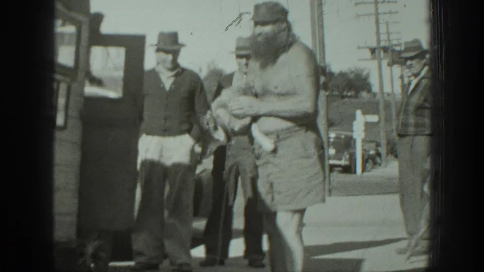A bearded man cradles a small dog on a California sidewalk in 1939 as several men stand near a bus or camper-like vehicle.