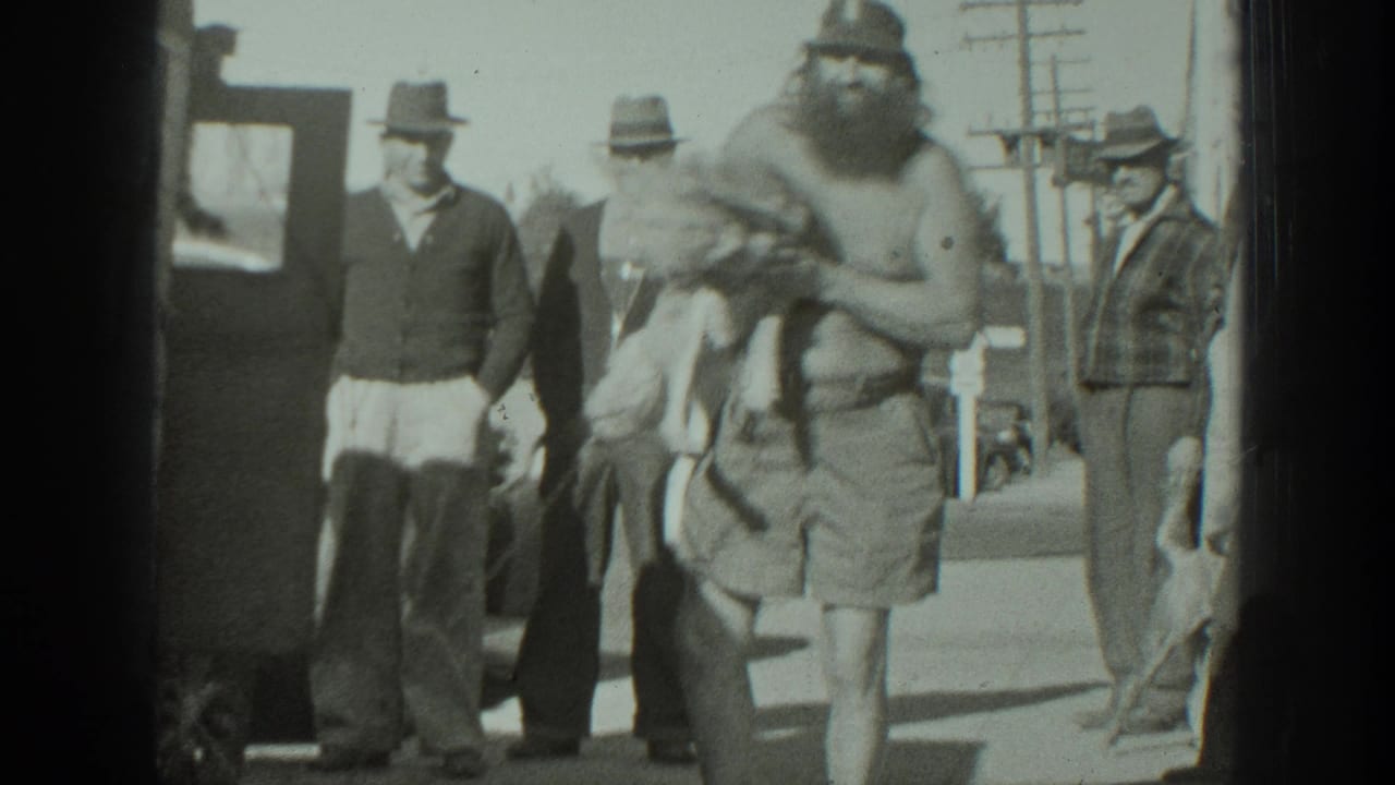 1939 California home-movie frame showing a bearded man carrying a small dog as several adult men in hats watch from the street.