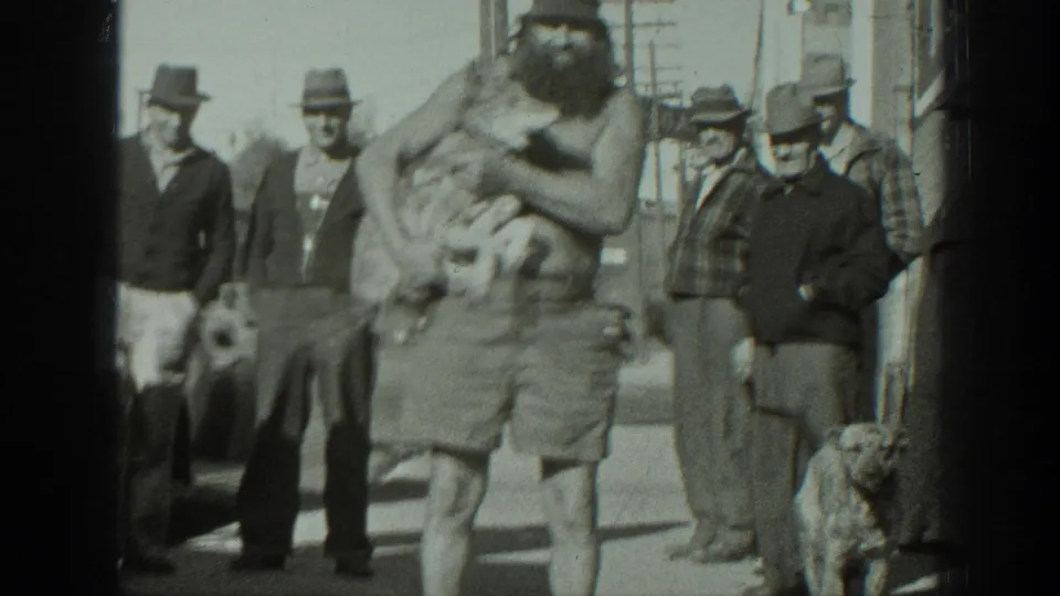 A bearded man in shorts holds a small dog close to his chest, with adult men in hats gathered behind him in 1939 California footage.