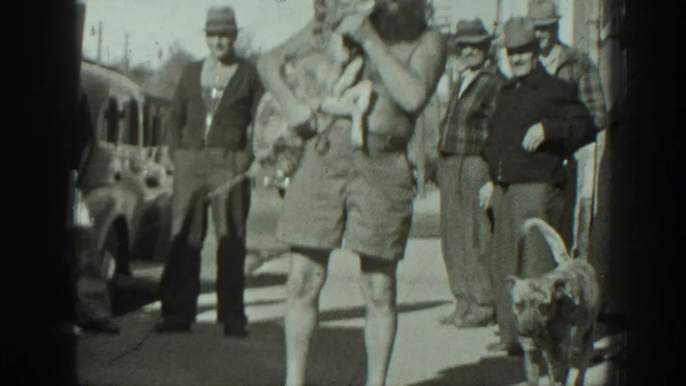 The bearded man carrying a small dog passes closer to the camera while another dog stands in the foreground near men in hats.