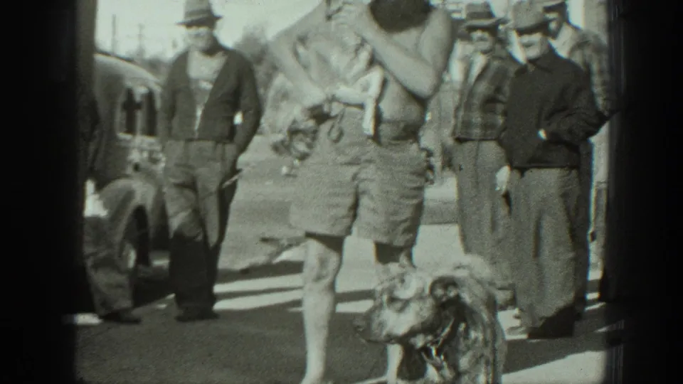 A dog stands near the camera while a bearded man carries another small dog past men in hats on a California street in 1939.