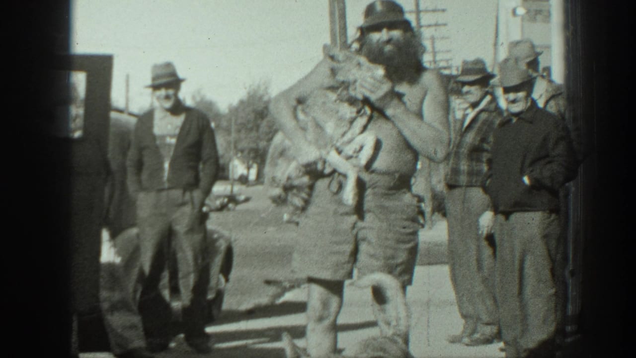 Shirtless bearded man holds two turtles on a street as adult onlookers and a dog stand nearby.