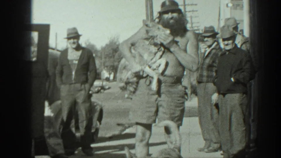 In 1939 California home-movie footage, a shirtless bearded man holds two turtles while adults watch on a street with a dog and cars nearby.