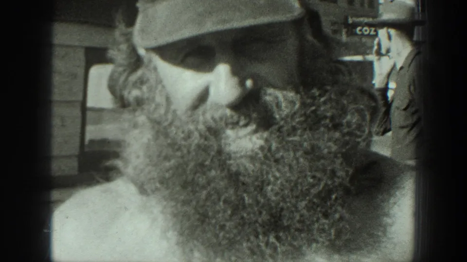 The bearded man in a cap is seen close up beside a tortoise, with outdoor structures faintly visible behind him.