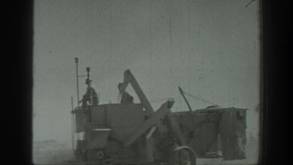 California 1939 frame showing a worker standing on a large grader-like machine, with metal arms and wheels visible.