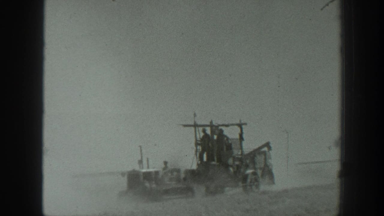 Large agricultural equipment travels across a field while people stand or ride aboard.